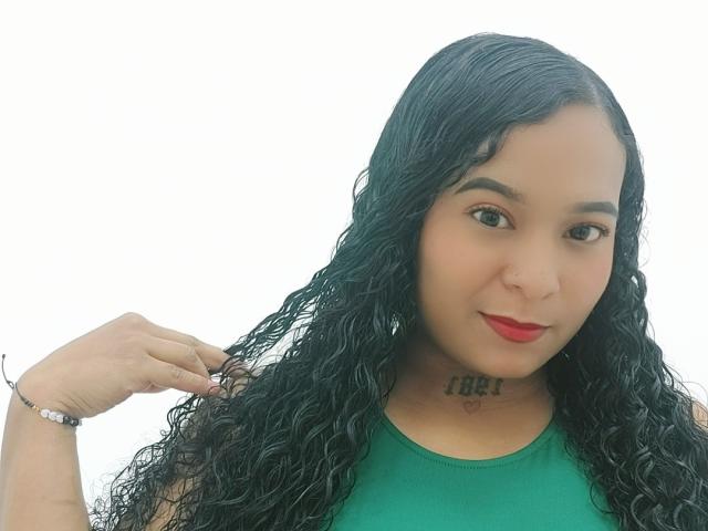 YirethHot met lang, krullend zwart haar, rode lippenstift, een groene top en een tatoeage onder de kin, die haar haar vasthoudt met een armband om de pols..