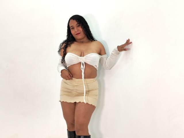 Yireth, avec ses cheveux noirs bouclés, un haut blanc à épaules dénudées qui révèle de grandes seins, une mini-jupe beige plissée, des bottes noires, debout devant un mur blanc, une main appuyée contre le mur, regardant l'appareil photo..