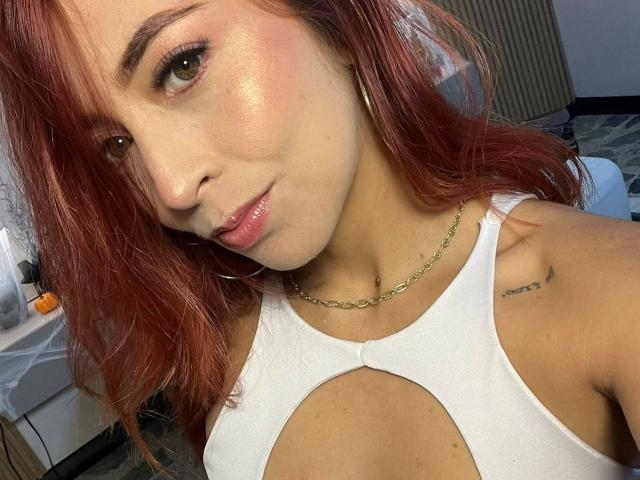 MiaaRouge with red hair, wearing a white cutout top, showing her tits, shiny pink lips, gold chain necklace, and small tattoo on her right shoulder.