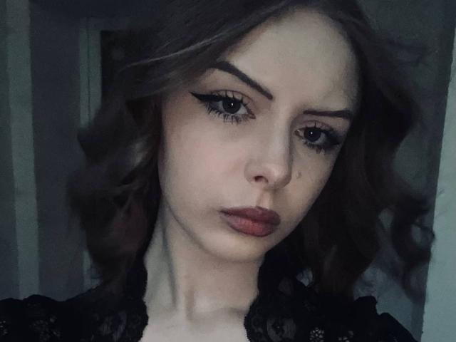 LourdesWhilby has wavy brown hair, thick eyebrows, black eyeliner, and red lipstick. She's wearing a black lace top, looking serious with a slight pout.