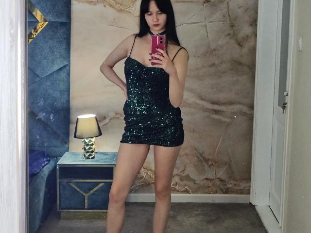 BerryDiva with dark hair, wearing a green sequin mini dress, holding a pink phone, standing in a room, showing her ass and legs, with a lamp on a table.
