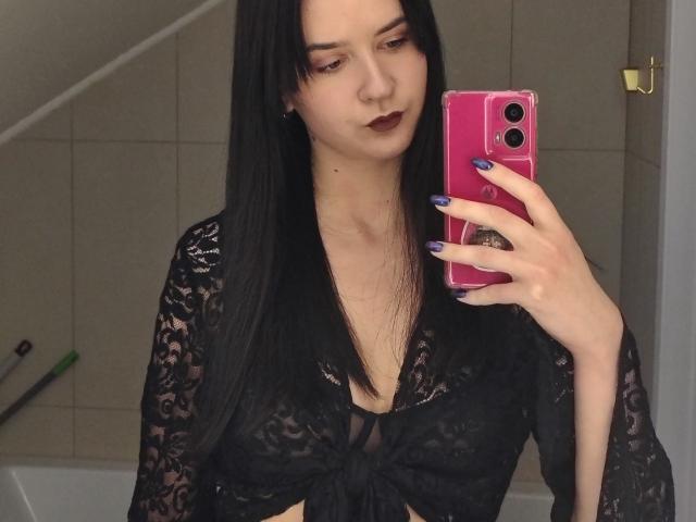 BerryDiva with long black hair, wearing a black lace top, holding a pink phone, showing her tits, dark lipstick, and purple nails, taking a mirror selfie.
