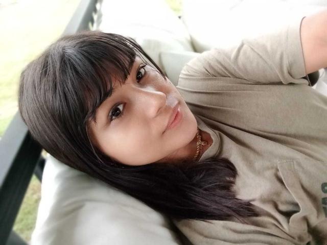 AshlyLee with straight black hair, bangs, brown eyes, and light skin, wearing a beige t-shirt, lying on a green cushion, looking up with a subtle smile.