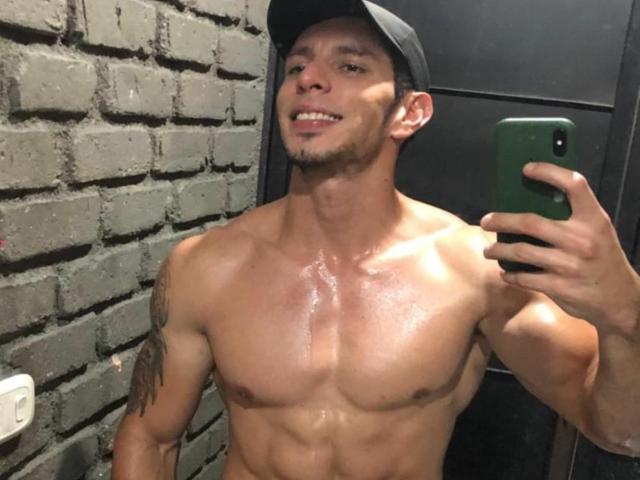 RobertHunk, shirtless with a sweaty, muscular torso, holding a phone in his right hand, wearing a black cap, smiling, and showing a tattoo on his left upper arm.