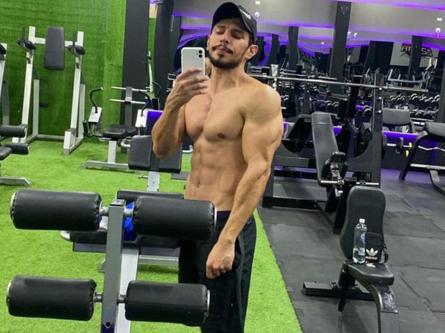 Muscular RobertHunk, shirtless with a defined six-pack, wearing black pants and a cap, taking a mirror selfie in a gym, showing off his strong arms and chest.