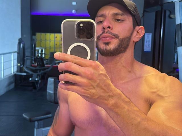 RobertHunk, shirtless, muscular, bearded, wearing a cap, holding a phone with a ring on it, taking a mirror selfie in a gym, showing his abs and tattoos.
