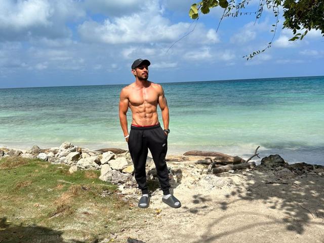Muscular RobertHunk stands shirtless, wearing black pants and a cap, with a bare chest and defined abs, hands in pockets, near a rocky beach, ocean in background.