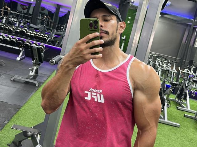 RobertHunk, muscular, sweaty, pink tank top, white trim, "Jordan" logo, black cap, holding green phone, mirror selfie, gym, green floor, visible biceps, short beard, focused expression.