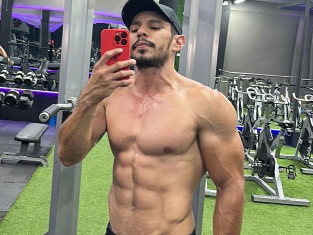 RobertHunk, shirtless, muscular, tan skin, defined abs, slight chest scars, black cap, holding red phone, taking mirror selfie, gym, green carpet, looking confident, slightly stubbled face.