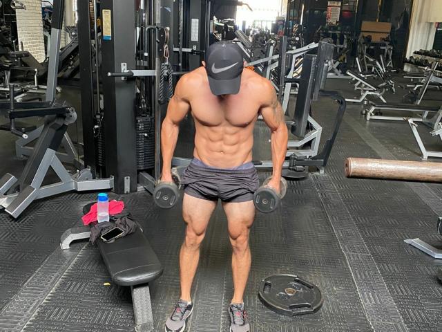 RobertHunk, muscular with defined abs, wearing a black Nike cap and gray shorts, stands in a gym, holding dumbbells, with a bottle and pink towel on a bench.