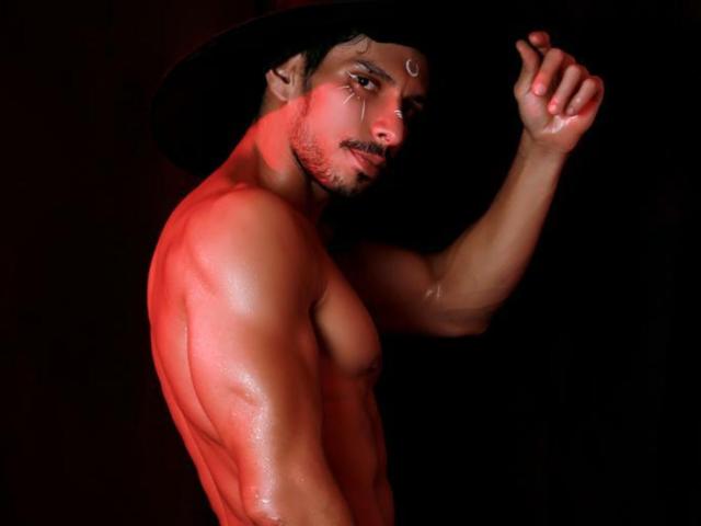 RobertHunk is a muscular dude with red-tinted skin, wearing a black hat, looking over his shoulder with a slight smirk, and his sweaty ass is prominently visible.