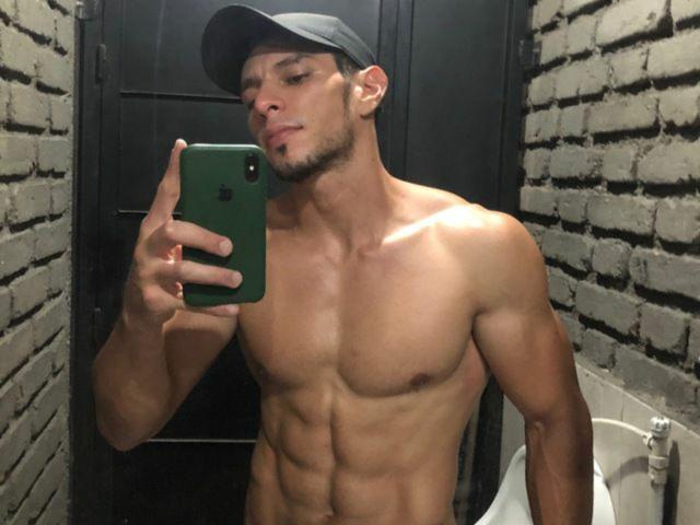 RobertHunk with a muscular, shirtless torso, wearing a black cap, holding a green phone in a mirror selfie, showing defined abs and a slight smirk.