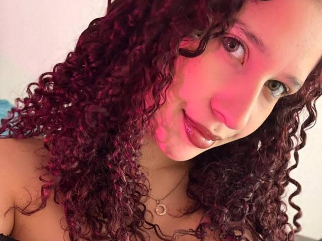 ValeryScot with curly dark red hair, medium skin, green eyes, shiny red lips, nose ring, and necklace, smiling at camera under pink lighting.