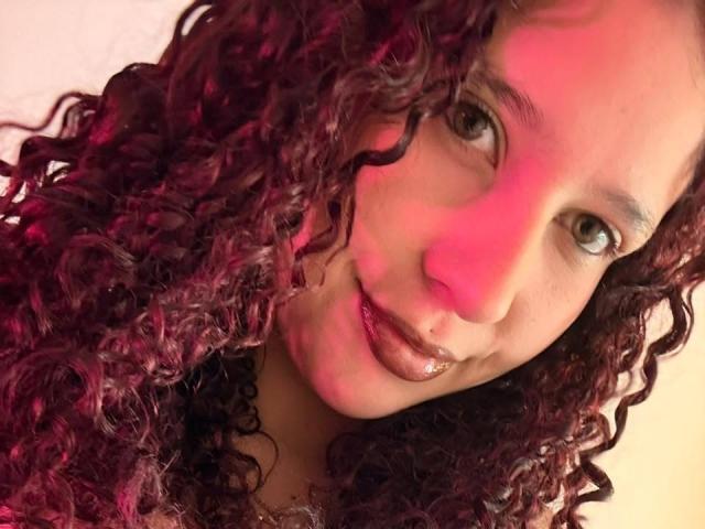ValeryScot with curly red-purple hair, glossy brown lips, and a cute smile, red light on face, green eyes, and a black choker, in a selfie.