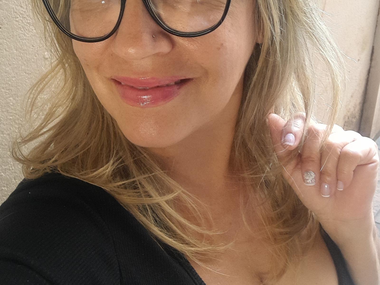Sheila69x mit glänzenden, rosafarbenen Lippen, schwarzen Brillen, blonden, gewellten Haaren, einem schwarzen Oberteil, die ihre rechte Hand mit einem Ring hält und ihre glänzenden Nägel zeigt. Ihre Brüste sind teilweise sichtbar..