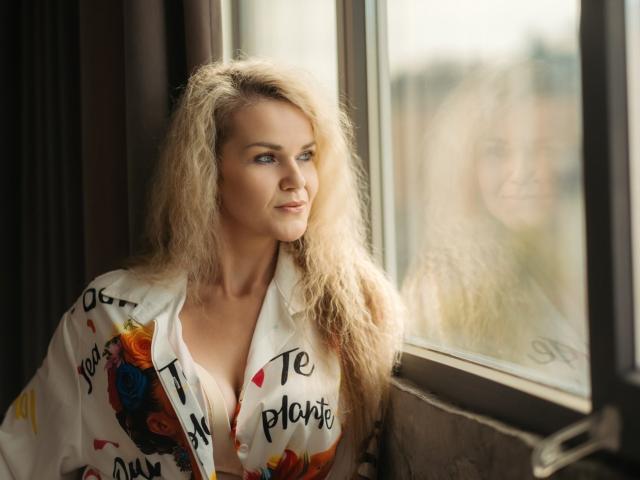 PenelopaMurMiay with curly blonde hair, looking out a window, wearing a colorful, partially open shirt, exposing her tits. Soft, natural light highlights her sexy, attractive face.
