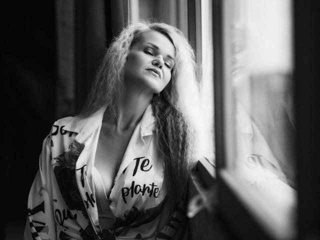 PenelopaMurMiay with long, curly hair, eyes closed, leaning against a window, wearing a loose shirt with floral and text patterns, sunlight casting a soft glow on her face.