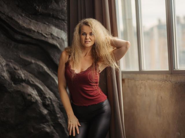 PenelopaMurMiay with long, wavy blonde hair, red tank top, and black leather pants, standing by a window, hand in hair, smiling subtly, looking sexy and confident.