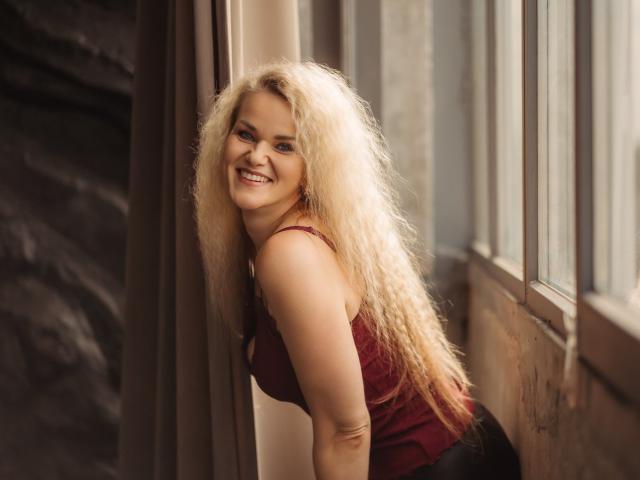 PenelopaMurMiay with long, curly blonde hair, smiling, wearing a red tank top, leaning against a window, showing her ass and a cute face, in a warm, natural light.