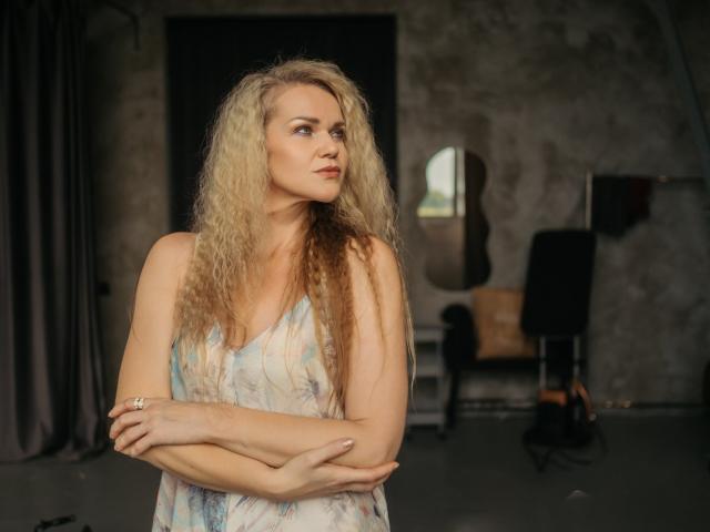 PenelopaMurMiay with long, curly blonde hair, crossed arms, and a white floral dress, looking to the side with a slight pout, in a dimly lit room.