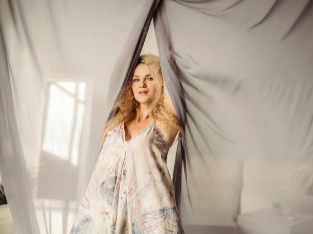 PenelopaMurMiay with blonde hair, wearing a floral dress, standing between semi-transparent gray curtains, smiling softly, sunlight casting gentle shadows on her face.