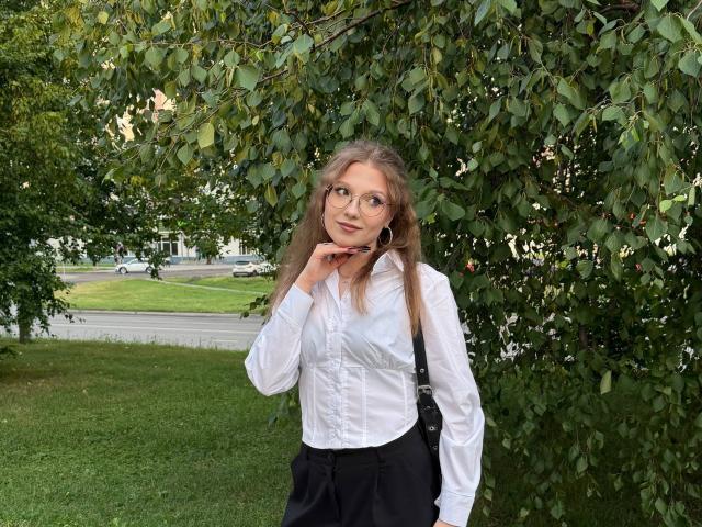Valrie Giuliano, con cabello rubio largo, gafas, camisa blanca, falda negra, mano en la barbilla, de pie debajo de un árbol de hojas verdes, bolso negro sobre el hombro, césped, carretera en el fondo..