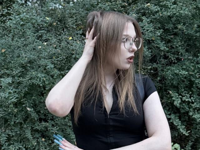 ValrieGiuliano, com cabelos castanhos longos, óculos, blusa preta, mão no cabelo, unhas azuis, olhando de lado, fundo com folhagem verde, pele clara, seios médios, maquiagem discreta, ambiente externo casual..