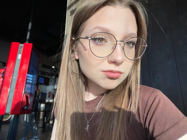 Valrie Giuliano, com cabelos castanhos longos, óculos redondos, piercing no nariz, brincos de argola e blusa marrom, olhando para a câmera, com luz do sol no rosto, batom rosa claro e colar com cruz..