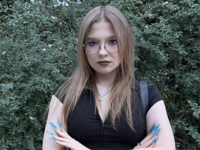 Valrie Giuliano, con cabello rubio largo, gafas, lápiz labial oscuro, camisa negra, brazos cruzados, uñas azules, collar, mochila negra, fondo con hojas verdes..