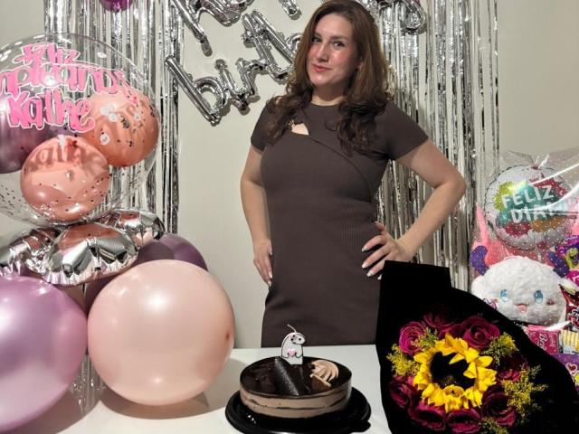 KatyaRogersX with brown hair in a brown dress, standing with hands on hips, next to a chocolate cake with a small white figure on top, and pink balloons.