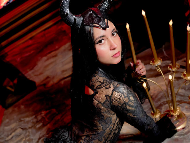 Gaby Mathew in einem schwarzen Spitzenoutfit, mit einem gehörnten Helm, Kerzen haltend, über die Schulter schauend, blasse Haut, langes schwarzes Haar, sexy, weiches Licht, Spitzenbesatz, Kerzen in der rechten Hand haltend..