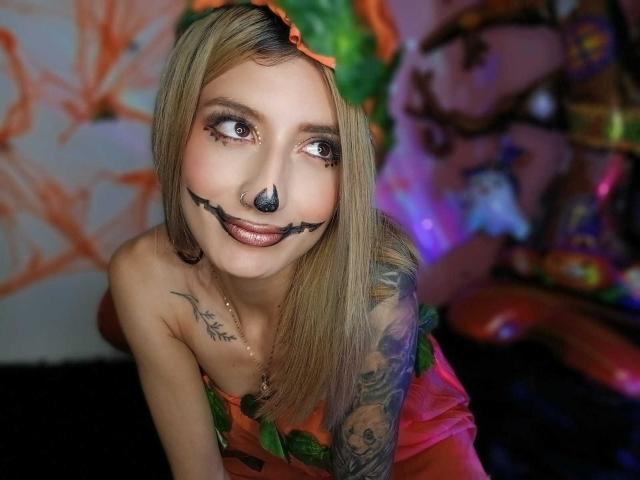 Kaiix met blond haar, donkere make-up op neus en lippen, een groene bladhoofdband, een oranje top zonder bandjes, een tatoeage op de rechterarm, leunt voorover, glimlacht, en heeft een zichtbare neusring..