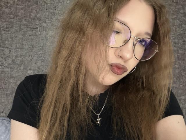 Valrie Giuliano, com cabelo castanho ondulado, óculos redondos, piercing no nariz e camiseta preta, franzindo levemente os lábios, usando um colar com uma cruz..
