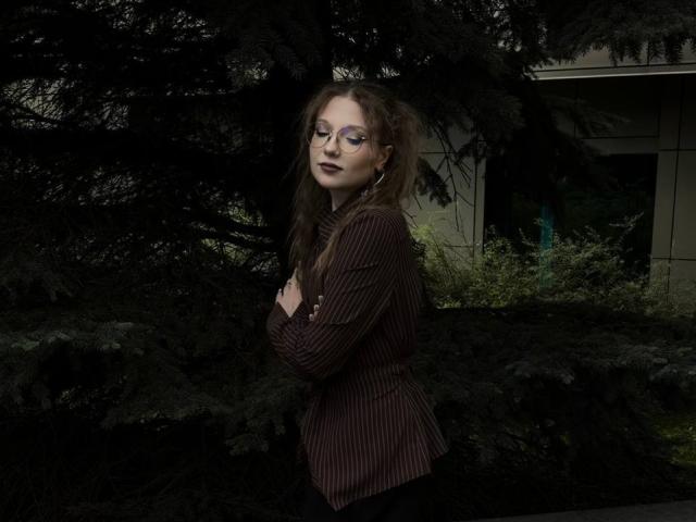 Valérie Giuliano mit Brille, dunklem Lippenstift und einem braun gestreiften Hemd, mit verschränkten Armen, steht vor einem dunkelgrünen Baum, schaut zur Seite, hat langes, braunes Haar und ein leichtes Lächeln..