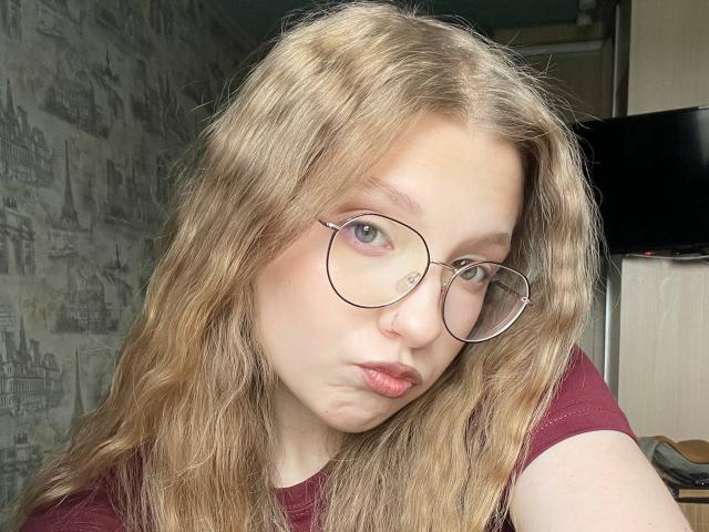 Valrie Giuliano, con cabello rubio ondulado, usa gafas redondas, tiene los labios fruncidos y viste una camisa color burdeos. Tiene la piel clara y mira directamente a la cámara. Tiene el cartílago de la nariz perforado..