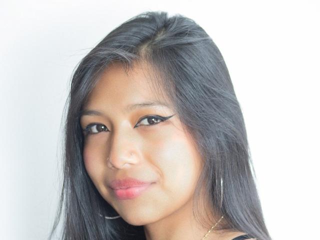 TianaCollins has black hair, a nose ring, and eyeliner with wings. She's smiling with pink lips, wearing a gold necklace, and has a slight tan.