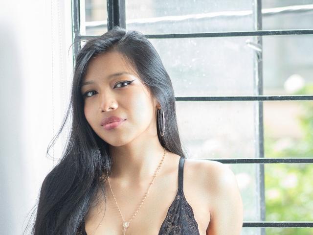 Tiana Collins, con cabello negro largo, vestida con un sujetador de encaje negro, con senos pequeños, piel clara y un collar dorado, está de pie junto a una ventana con barrotes de metal..