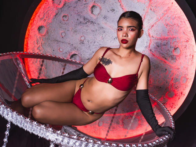 TaylorGil with dark skin, red lingerie, black gloves, and red lipstick, sitting in a silver, pearl-embellished chair, showing small tits, tattoo on right chest, and a confident look.