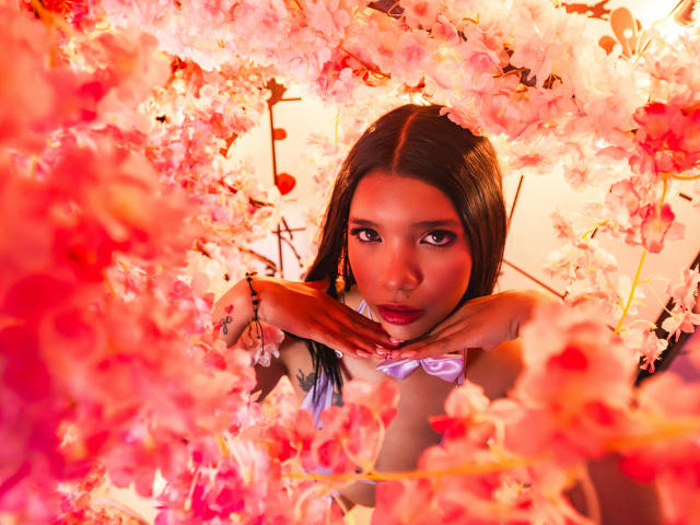 TaylorGil with dark brown hair, pink floral frame, hands on face, purple lipstick, gold earrings, looking seductively at camera, surrounded by pink cherry blossoms.