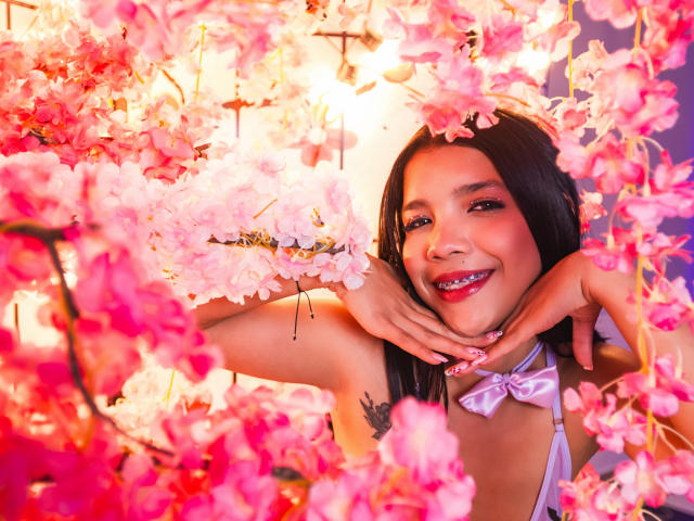 TaylorGil with pink cherry blossoms, smiling with pink lipstick, hands up, showing a tattoo on her right arm, wearing a pink bow, in a bright, colorful, floral setting.