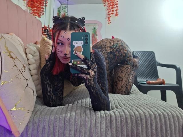 AnaRossii with red hair, black cat ears, and facial paint, wearing a lace bodysuit, on a ribbed bed, taking a selfie, showing her butt and fishnet legs.