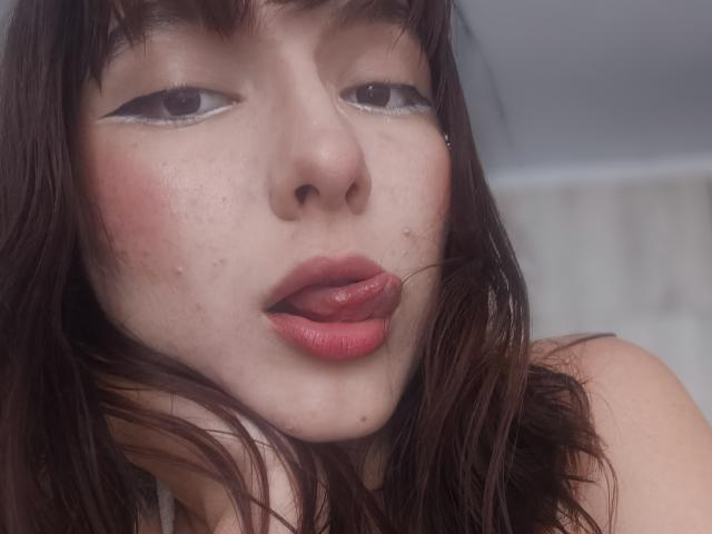 ValeriHairy69's face, showing pinkish cheeks, brown wavy hair, and red lips with a small bit of her tongue sticking out. She has eyeliner, freckles, and a nose piercing.