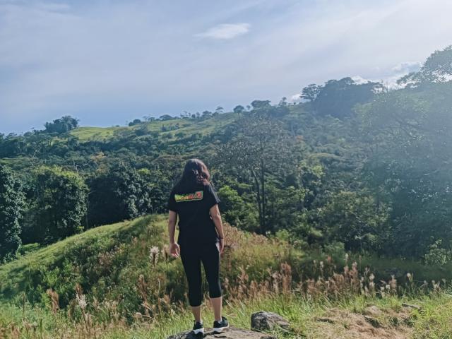 ValeriHairy69 stands with back to camera, wearing black shirt and capri pants, in sunlit green hill landscape, grassy foreground, rocky path, and blue sky with light clouds.
