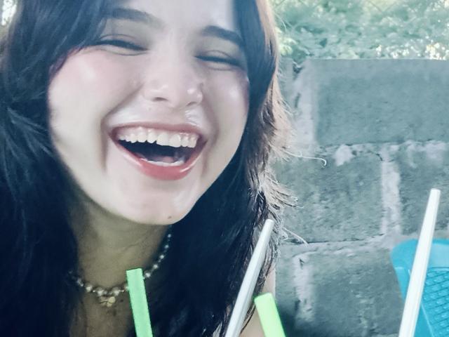 ValeriHairy69 with a big, open-mouthed smile, eyes closed, holding green and white straws, wearing a necklace, and has dark hair and fair skin.