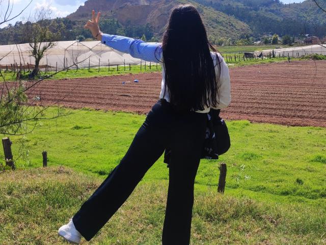 ValeriHairy69 with long black hair, blue shirt, black pants, white socks, and white shoes, makes a peace sign, standing on green grass with a raised leg, facing farmland.