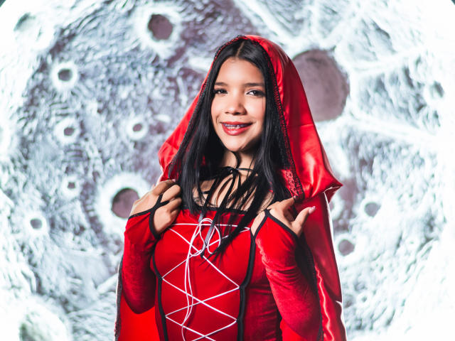 TaylorGil with long black hair, red hooded dress, and lace-up bodice, smiling, holding hood strings, showing small tits, cute, wearing red lipstick, looking sexy.