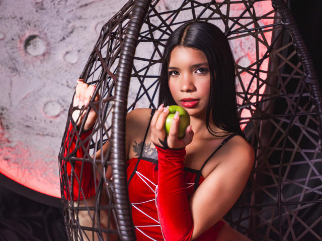 TaylorGil with dark hair, red velvet top, and matching arm sleeves, holding a green apple in a wicker chair, looking at the camera with a serious expression.
