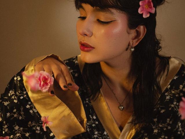 ValeriHairy69 with dark hair, pink flower, gold-lined black floral robe, red lipstick, shiny eyes, black nail polish, holding pink flower, wearing heart necklace, soft brown background.