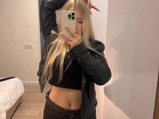 DelphiaCabos with blonde hair, black leather jacket, black crop top, and brown pants, taking a mirror selfie with her left arm raised, exposing her flat, toned midriff.