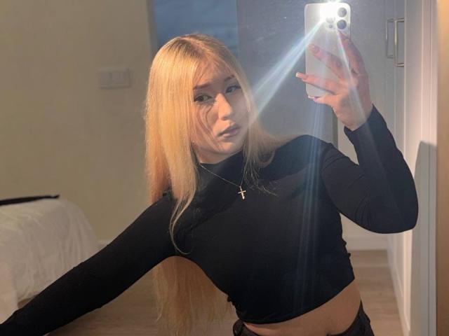 DelphiaCabos with long blonde hair, black long-sleeve top, cross necklace, holding phone, mirror selfie, light reflection, slight pout, midriff visible, bed in background.